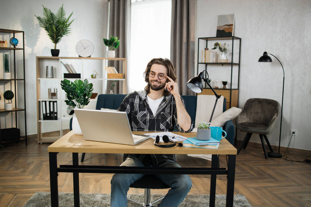 Bearded young man in checkered shirt and eyeglasses sitting at table with modern laptop and writing notes in modern office. Concept of freelance and technology. - Valokuva, kuva