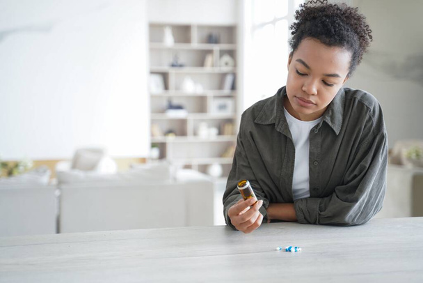 Unwell african american girl is holding pills flacon. Sick teenage girl is taking vitamins or painkillers. Young woman is upset and worried. Stressed patient. Pharmaceutical treatment concept. - Fotoğraf, Görsel