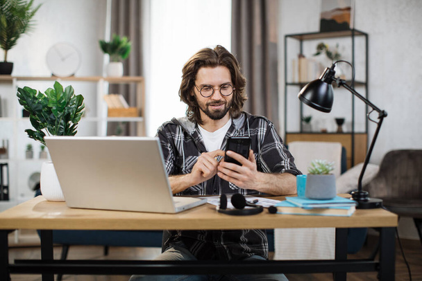 Young bearded excited man in casual outfit typing on mobile phone while sitting at workplace with laptop. Concept of corporate communication and good news. - Photo, image