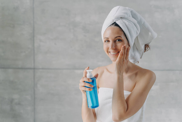 Face toner or micellar water advertising concept. Happy young woman touches pure skin. Gorgeous caucasian woman wrapped in towel after bathing applying toner in flacon. Face washing foam in bottle. - Foto, Bild
