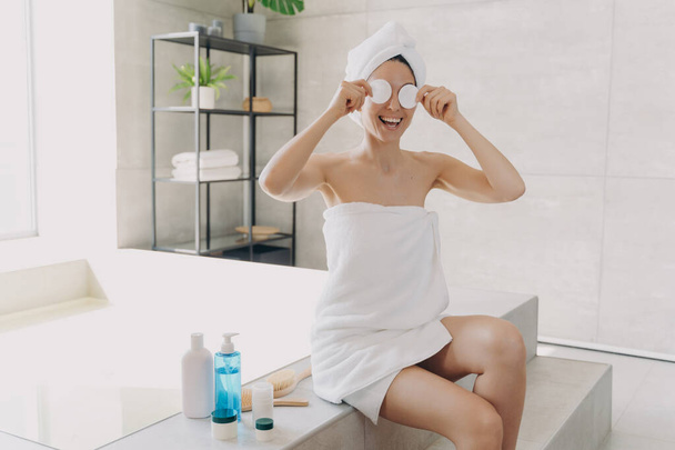 Happy young woman has fun applying face toner with cotton discs. Gorgeous european girl wrapped in towel after bathing. Lady is sitting on bathtub and relaxing. Woman takes shower at home. - Foto, Imagem