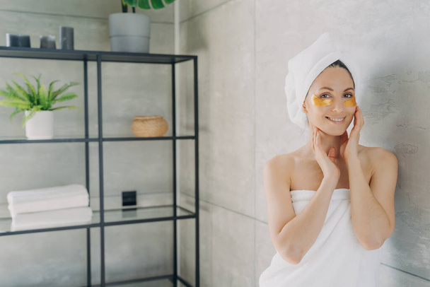 Relaxation at spa resort. Happy girl wrapped with her hair and body in towels applies eye patches. Young hispanic lady takes shower at home. Face skin rejuvenation and collagen nourishing. - Фото, зображення