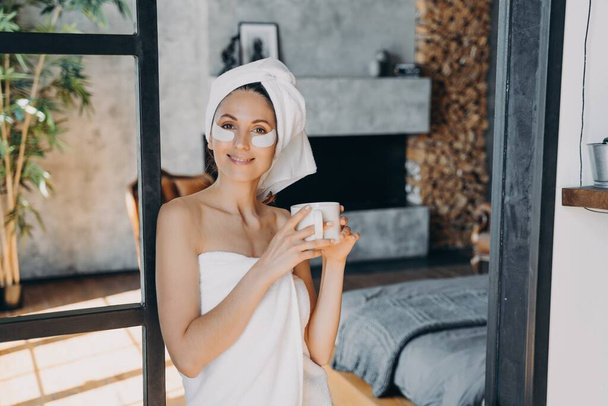 Girl applies lifting eye patches. Weekend morning beauty routine at home Young woman with cup of tea relaxing at luxurious spa resort. Attractive european woman wrapped in towel after bathing. - Foto, imagen