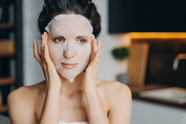 Caucasian girl applies mask sheet after bathing. Vitamin, nourishing and nutrition face mask. Young woman takes shower at home or hotel and doing spa procedures. Skin care and rejuvenation. - 写真・画像