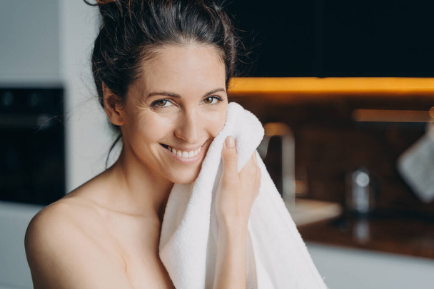 Caucasian woman is wiping face with towel after washing and smiling. Young woman takes shower at home and doing daily skin care. Hygiene and wellness, dermatology and skin cleansing. - Foto, imagen