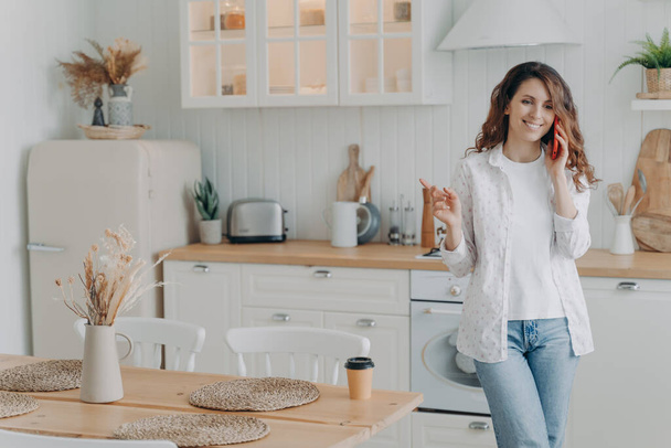 Νεαρή Ευρωπαία γυναίκα μιλάει στο smartphone της στην κομψή κουζίνα της στο σπίτι. Ευτυχισμένη ελκυστική νοικοκυρά έχει ελεύθερο χρόνο. Μοντέρνο λευκό σκανδιναβικό εσωτερικό. Κουζίνα, πάγκος εργασίας και κουζίνα. - Φωτογραφία, εικόνα