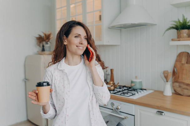 Young caucasian woman with phone has conversation in her stylish kitchen at home. Morning coffee at modern white scandinavian interior. Stove, worktop and cuisine. - Photo, Image