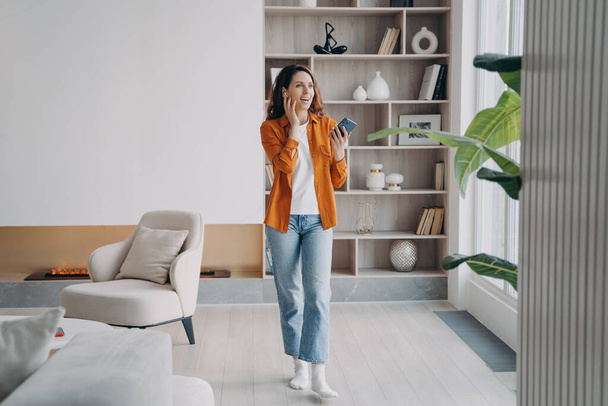 Happy european girl has phone call at home. Pretty young woman is using airpods. Wireless earphones. Technology using, leisure and communication concept. Beautiful modern interior. - Φωτογραφία, εικόνα