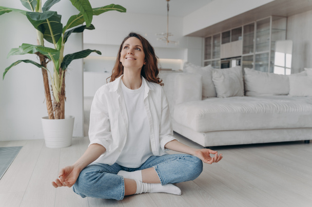 Peace of mind and mental health concept. Young european woman is practicing yoga on floor and smiling. Meditation, consciousness and relaxation at home. Posture exercise, lotus asana. - Foto, Imagem