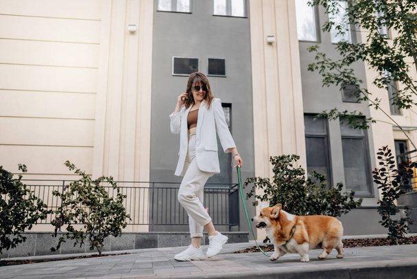 Женщина в белом костюме и солнцезащитных очках прогуливается по улице с валлийской собакой Корджи Пемброу - Фото, изображение