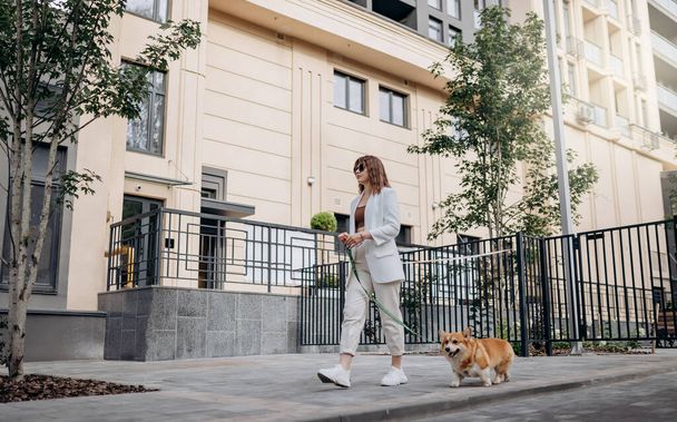 Женщина в белом костюме и солнцезащитных очках прогуливается по улице с валлийской собакой Корджи Пемброу - Фото, изображение
