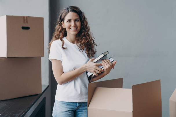 Happy european woman unpacking boxes and taking off books and notes. Young student is housing in campus room. Girl in white t-shirt indoors. Relocation and independence concept. - Zdjęcie, obraz