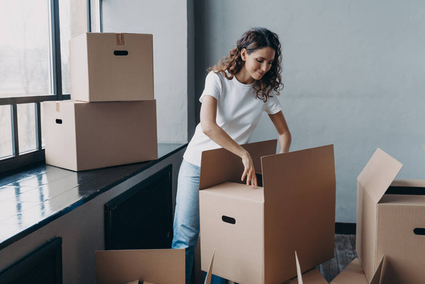 Happy young european woman unpacking boxes. Girl near window in white t-shirt indoors. Woman is opening cardboard box and smiling. Relocation and independence concept. - Zdjęcie, obraz