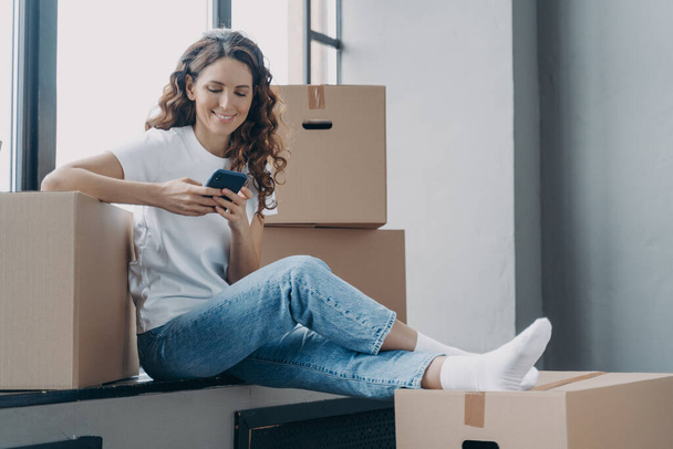 Happy spanish woman is sitting on windowsill of new home. Girl is unpacking boxes. Young lady is texting on phone and smiling. Joy of relocation concept. - Foto, Imagem