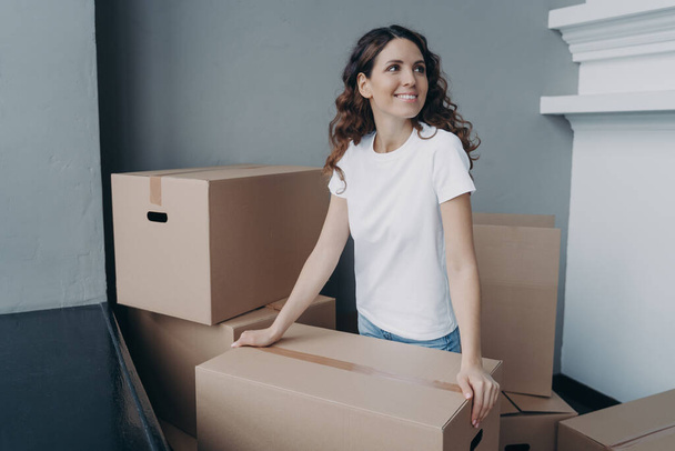 Hopeful lady is holding cardboard box and moving to modern apartment. Happy european woman purchasing real estate and looking at interior. Improvement, changes and new life concept. - Foto, Bild