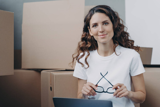 Girl is sitting on floor with laptop in new apartment. Happy hispanic woman in glasses with pc among cardboard boxes. Distance work from any place and freelance concept. - Foto, imagen