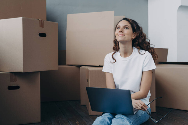 Hopeful hispanic woman sitting among cardboard boxes and holding pc. Lady is in waiting for shipping service and chatting online. Romantic relationship and relocation concept. - Foto, Bild
