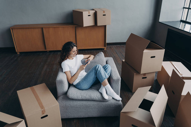 Girl having rest in new apartment and clicks phone. Successful independent spanish woman is moving. Living room interior and cardboard boxes. Mortgage loan and dream house purchase concept. - Foto, Bild