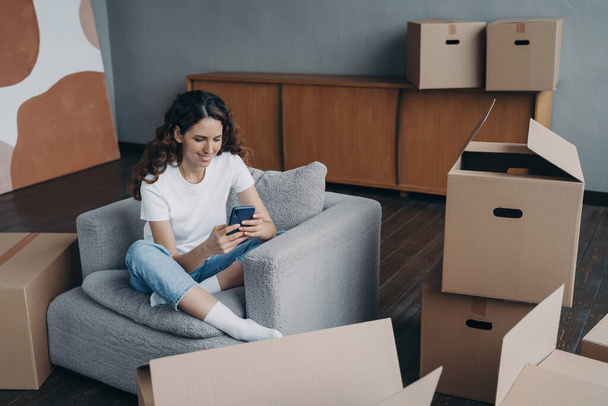 Young spanish woman posting news. Happy apartment buyer unpacking boxes. Girl is moving to new home. Relaxed young lady clicks mobile phone. Independence and achievement concept. - Zdjęcie, obraz
