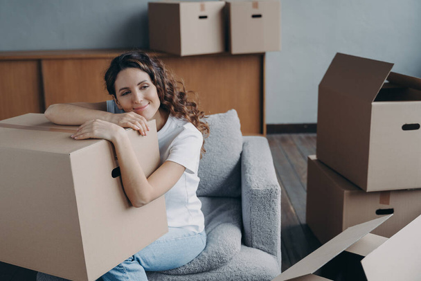 Hispanic girl relocates to new home. Lady is sitting in armchair among packed cardboard boxes. Woman is dreaming leaned to box. Happy young woman moves alone. Independence and success concept. - Zdjęcie, obraz
