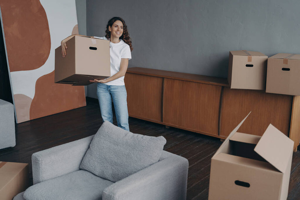 Happy young woman relocates alone and carrying a box. Single spanish lady moves. Apartment selling. Real estate purchase, mortgage, delivery service ordering concept. - Photo, Image