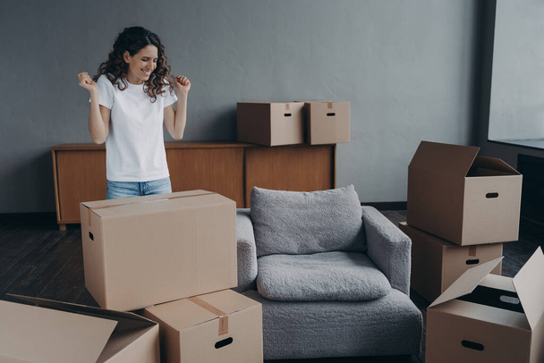 Excited young woman is packing things. Spanish girl moves to new house. Lady is unpacking cardboard boxes in new apartment. Big new living room with furniture. Real estate purchase concept. - Photo, image