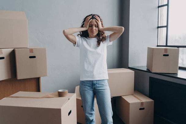 Headache from moving. Exhausted woman is tired from packing cardboard boxes with duct tape. European young lady in jeans and white t-shirt going to relocate. Anxiety and stress from moving concept. - Valokuva, kuva