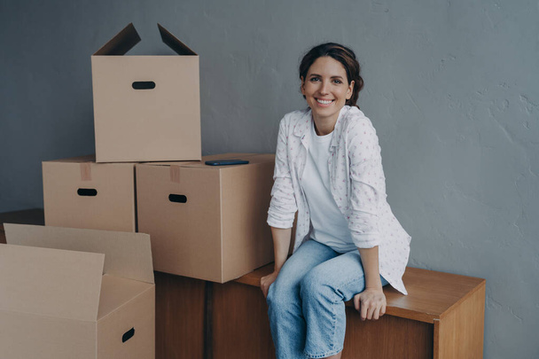 Happy teenage girl moves. Hispanic lady is sitting on boxes in new apartment and smiling. Attractive young woman unloading containers. Concept of success and opportunities for women. - Fotoğraf, Görsel