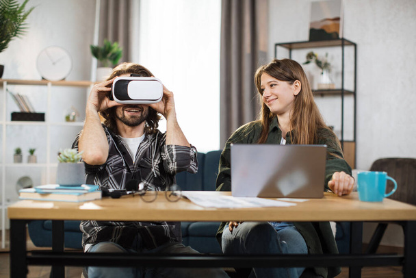 Excited young people with surprised expression, playing games at home and using VR glasses and laptop. Happy man wears VR headset, while pretty girl uses laptop and smiles - Фото, зображення