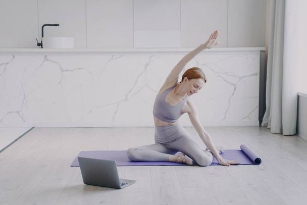 Young happy white woman is stretching and practicing yoga on mat. Concept of e-learning and home online video classes. Laptop is on floor. Sport online and healthy lifestyle on quarantine. - Fotoğraf, Görsel