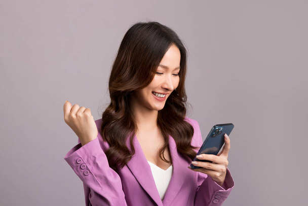 Азиатка с длинными волосами, в розовом костюме, держит смартфон, счастливые жесты на сером фоне. - Фото, изображение