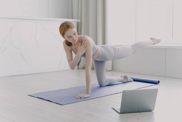 Female coach explaining at video class. Young athlete practicing yoga by video lessons in her apartment. White woman doing posture exercise. Concept of personal training and remote work. - Fotoğraf, Görsel