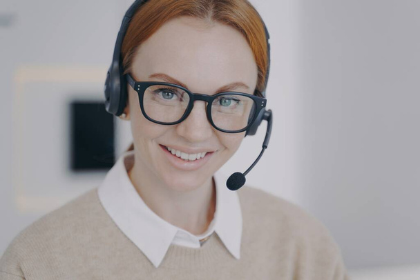 Young woman in earphones sitting at workplace. Online consultant of call center. Caucasian friendly girl in headset provides information by telephone. Customer support concept. - Fotoğraf, Görsel