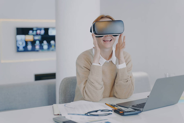 Young european woman in VR headset in office. Excited girl student is sitting at the desk in front of laptop. Concept of interactive innovation and futuristic university. Modern device using. - Foto, imagen