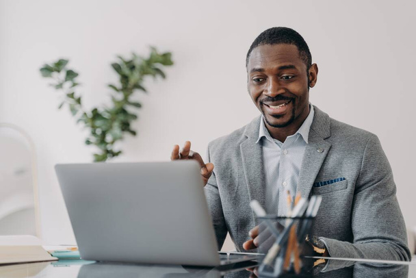 Young man has video conference on laptop. Freelancer is working online at workplace at home and talking to client. Remote african american worker is suit is sitting in front of camera. - Foto, Imagen