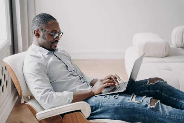Διαδικτυακή ανεξάρτητη δουλειά. Όμορφος Αφροαμερικάνος μπροστά στον υπολογιστή. Χαλαρωμένος χαρούμενος επιχειρηματίας με επίσημα ρούχα κάθεται σε αναπαυτική καρέκλα στο διαμέρισμά του. Έργο εκκίνησης, απομακρυσμένη εργασία. - Φωτογραφία, εικόνα