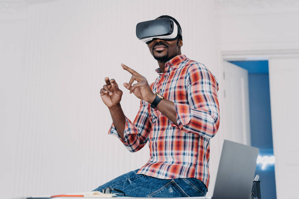 Happy african american guy in vr goggles at home. Young businessman has virtual study. Executive is working on futuristic design project. Modern digital gadget for business and e-learning. - Foto, Imagem