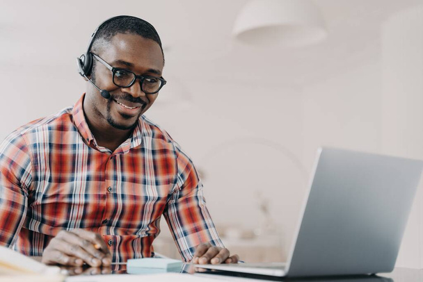 Χαμογελώντας Αφροαμερικανός υπάλληλος που φοράει ακουστικά δουλεύοντας στο λάπτοπ, κοιτάζοντας την οθόνη, απαντώντας σε μια βιντεοκλήση, επικοινωνεί με τον πελάτη. Μαύρος τηλεφωνητής κέντρο παροχής συμβουλών πελάτη. - Φωτογραφία, εικόνα