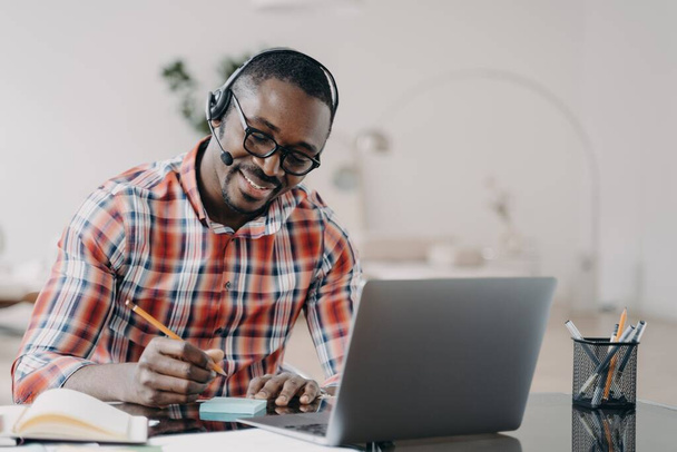 African american student young man getting remote education at laptop. Happy black male wearing headset listening lecture, taking notes while online lesson by video conference. Distance learning  - Fotografie, Obrázek