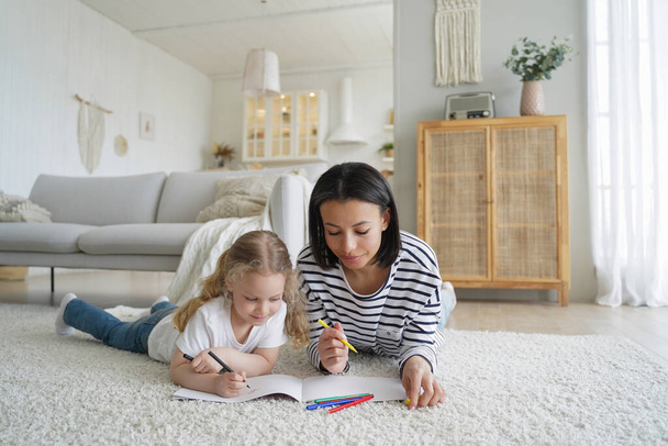 Mom and her kid are drawing together. Young caucasian woman and little daughter lying on floor together and coloring with markers. Happy family has good time at home. Childhood and parenthood concept. - Foto, Imagem