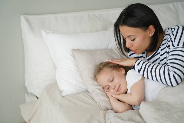 Mom puts daughter to bed, stroke head of small child during daytime nap. Calm kid girl rest sleeping lying on soft pillow under fresh duvet. Healthy sleep in children, childcare. - Zdjęcie, obraz