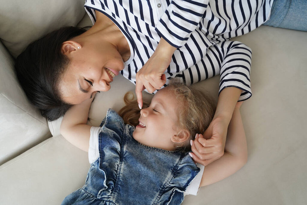 Нежность к ребенку. Молодая мама наслаждается утром со своей маленькой дочерью на диване. Кавказская семья играет и отдыхает вместе. Женщина и ребенок улыбаются и просыпаются после дневного сна. - Фото, изображение