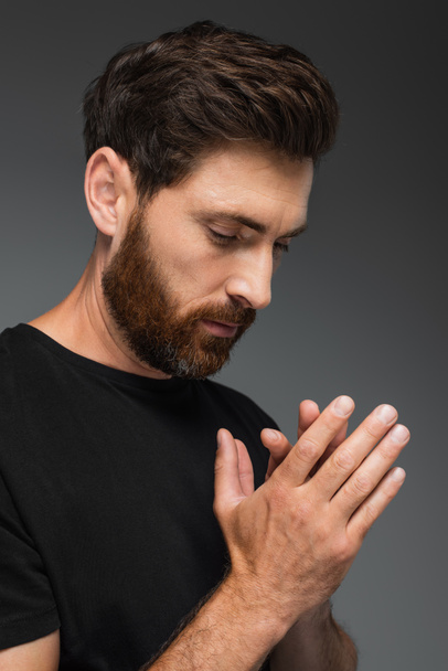 πορτρέτο του γενειοφόρου άνδρα σε μαύρο t-shirt προσεύχεται απομονωμένο σε γκρι - Φωτογραφία, εικόνα