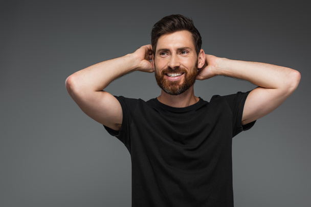 portrait of cheerful man in black t-shirt posing with hands behind head isolated on grey - Photo, Image