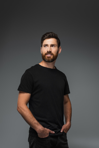 portrait of pleased bearded man in black t-shirt posing with hands in pockets isolated on grey - Foto, immagini
