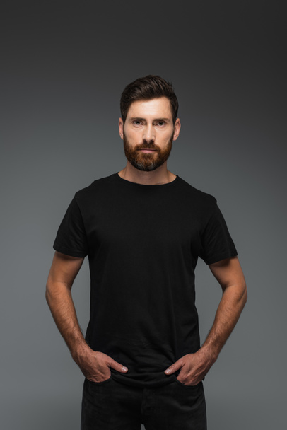 portrait of bearded man in black t-shirt looking at camera while posing with hands in pockets isolated on grey - Foto, Imagem