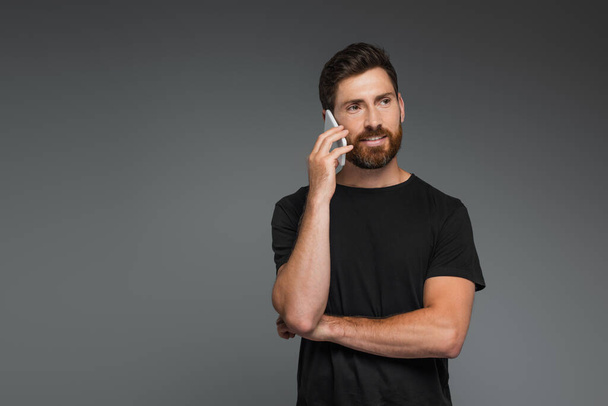 positive and bearded man in black t-shirt talking on smartphone isolated on grey - Foto, imagen
