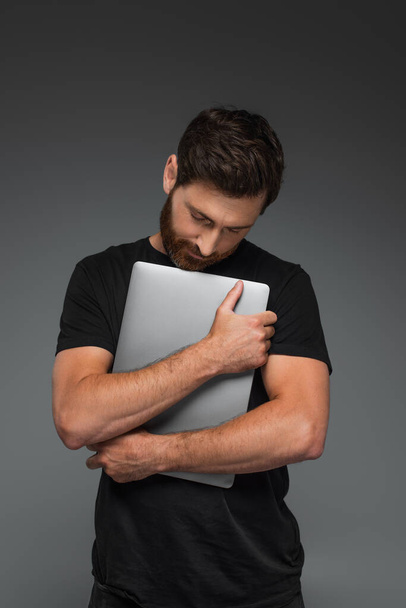 bearded man in black t-shirt hugging laptop isolated on grey - Foto, afbeelding