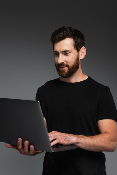cheerful and bearded man in black t-shirt using laptop isolated on grey - Fotó, kép
