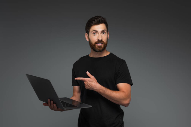 amazed and bearded man in black t-shirt pointing with finger at laptop isolated on grey - 写真・画像
